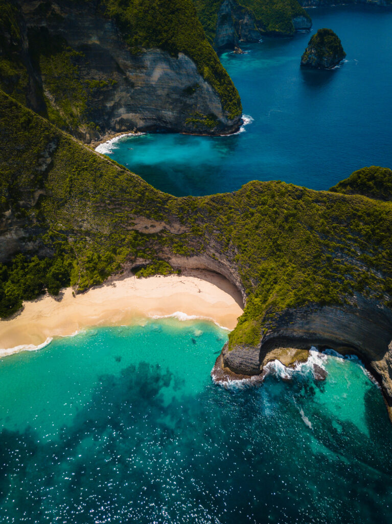 Pemandangan udara pantai berpasir putih dengan air laut biru kehijauan yang dikelilingi tebing hijau menjulang.