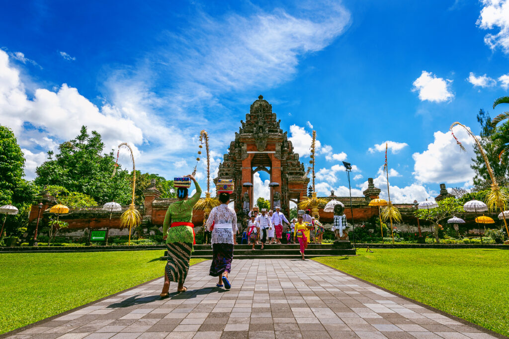 Umat Hindu Bali berjalan menuju pura saat upacara, mengenakan pakaian adat tradisional di bawah langit biru cerah.