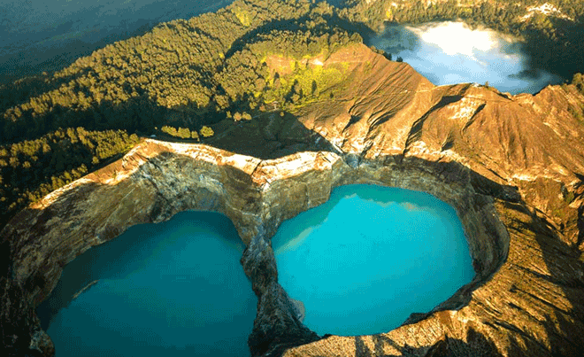 Tiga kawah berisi danau berwarna cerah di puncak Gunung Kelimutu, dikelilingi hutan hijau dan kabut tipis.