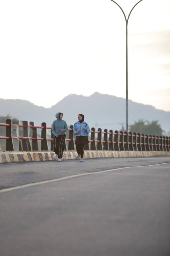 Dua perempuan berlari santai di jalan raya dengan latar pegunungan saat pagi hari, mencerminkan tren running di media sosial